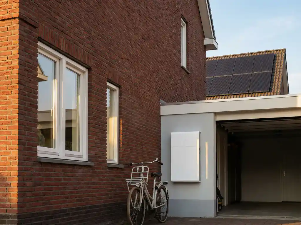 Witte Sigenergy thuisbatterij gemonteerd op garagemuur naast zonnepanelen in Nederlandse woonwijk met rode bakstenen en fiets